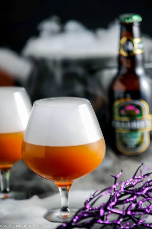 Orange Alcoholic Butterbeer Recipe in snifters with white fog on top, Halloween decor in background.