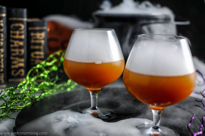 Bourbon Butterbeer Cocktail - orange cocktails in brandy snifters with dry ice clouds on top. Green glitter branches and black cauldron with dry ice behind. Gold print black books