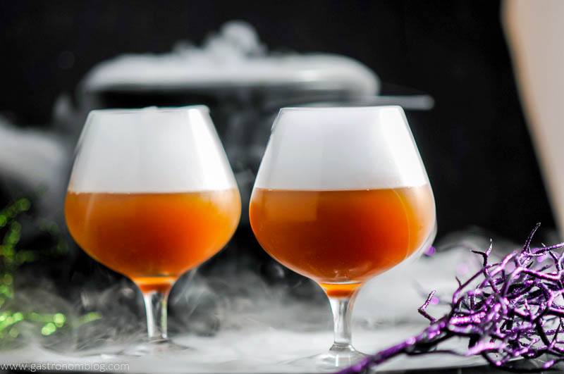 Orange Alcoholic Butterbeer Recipe in snifters with white fog on top, Halloween decor in background.