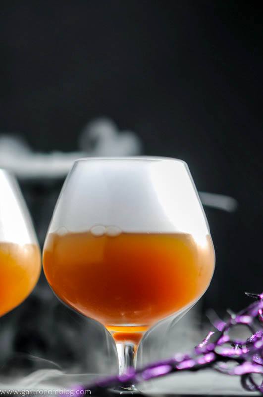 Orange Alcoholic Butterbeer Recipe in snifters with white fog on top, Halloween decor in background.