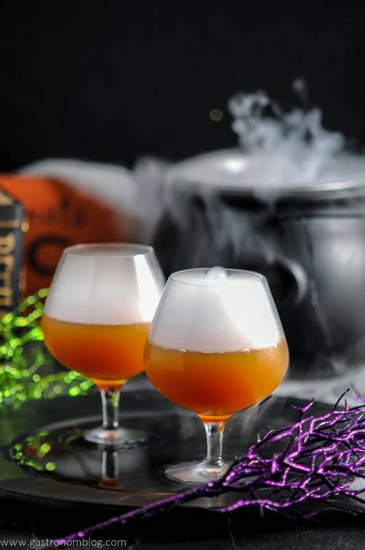 Bourbon Butterbeer Cocktail - orange cocktail in snifters with dry ice cloud on black plate, purple and green glitter stems. Black cauldron with dry ice behind
