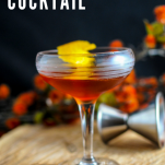whiskey cocktail in coupe with orange peel, on a wood board. JIgger and flower in background