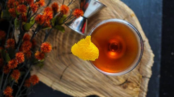 The Ginger Snap, brown whisky cocktail in coupe with lemon peel, top shot on wood board with jigger and autumn flowers
