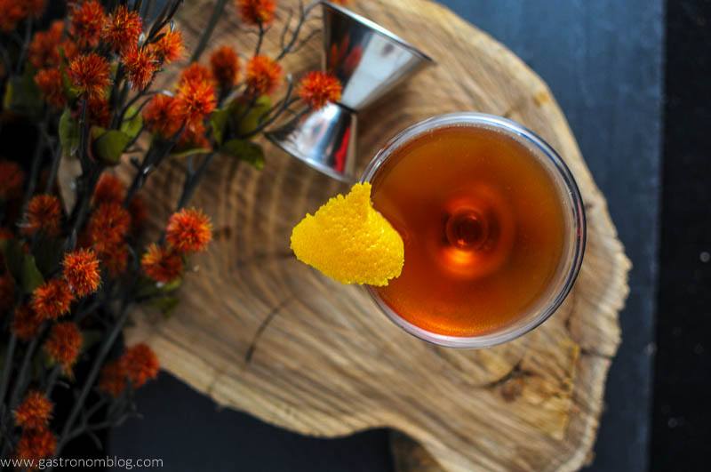 The Ginger Snap cocktail in cocktail coupe with lemon peel. JIgger and flowers on wood plate