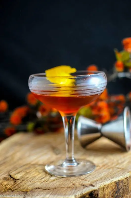 The Ginger Snap, brown cocktail in coupe with lemon peel on wood board, jigger and autumn leaves