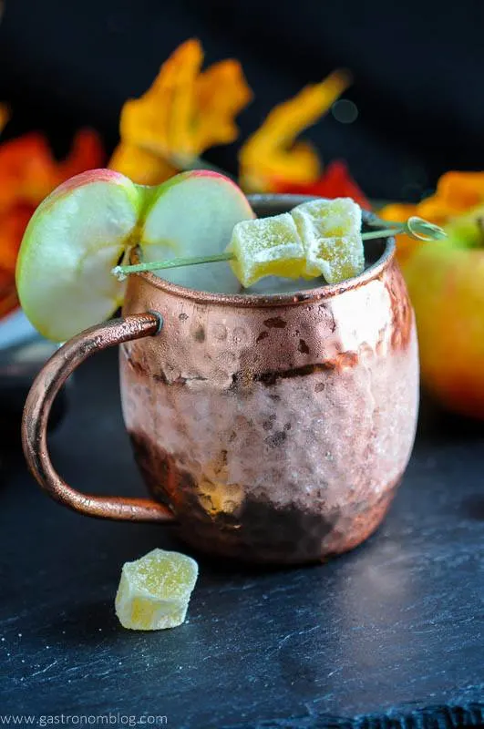 The Maple and Cider Kentucky Mule in copper mug with apple slice and candied ginger, autumn leaves behind