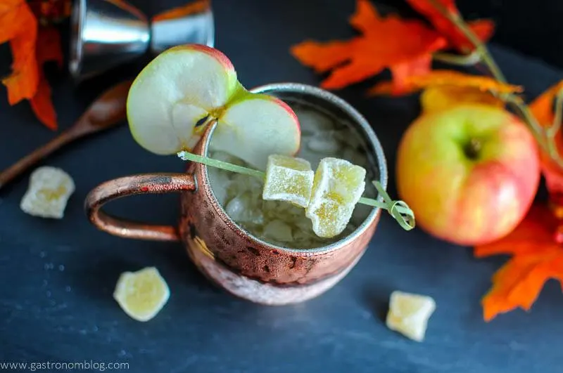 The Maple and Cider Kentucky Mule in copper mug. Apple slice and candied ginger. Wooden spoon, jigger, apple and fall leaves in background