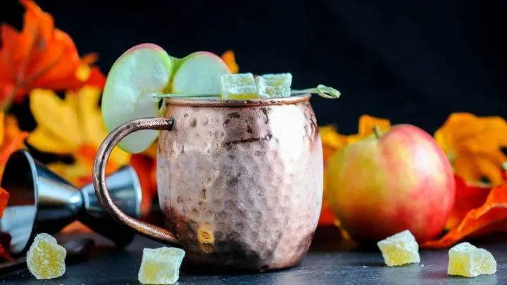 The Maple and Cider Kentucky Mule in a copper mug with apple slice and candied ginger, autumn leaves and apple behind
