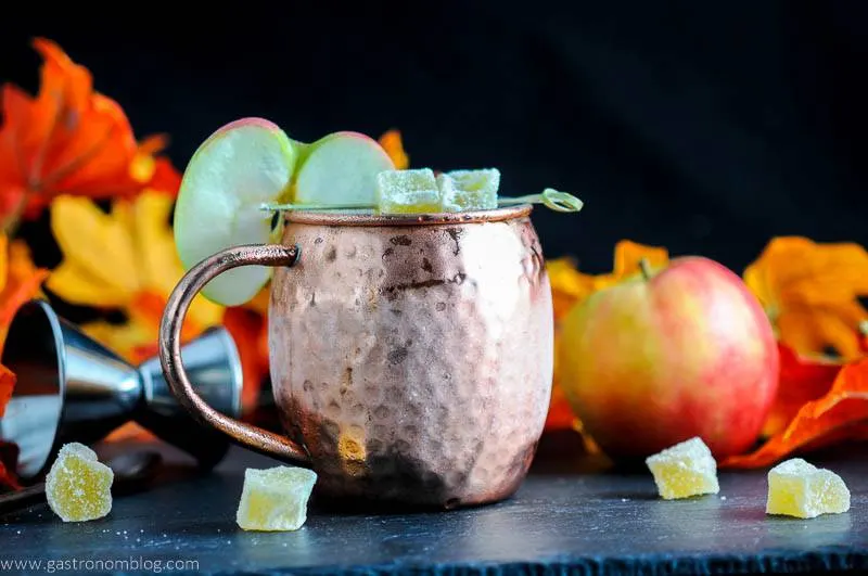 The Maple and Cider Kentucky Mule in copper mug. Apple slice and candied ginger, jigger, apple and fall leaves in background