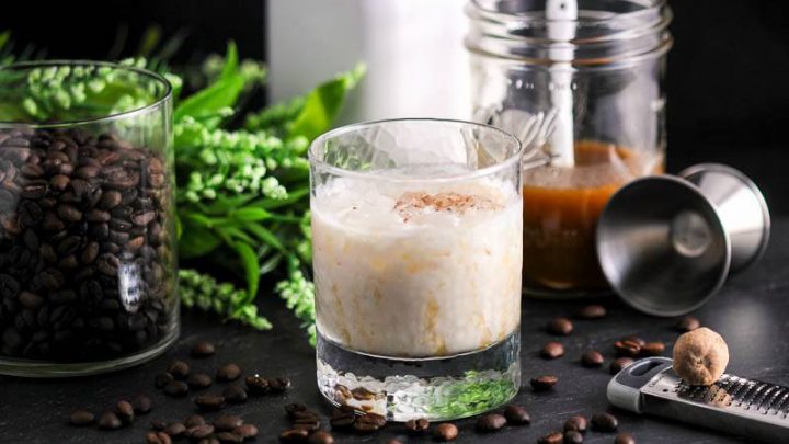 Salted Bourbon Butterscotch White Russian Cocktail in rocks glass. Greenery, white bottle, jar of butterscotch, jigger and coffee beans around glass