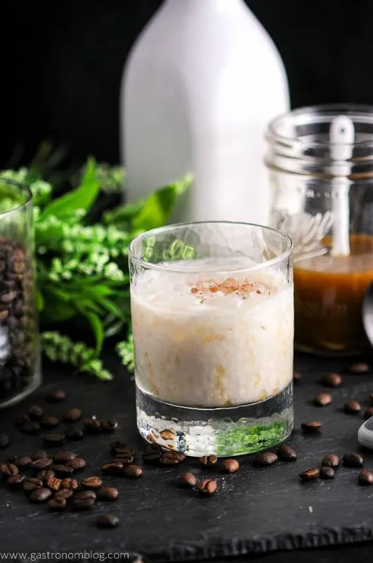 Salted Bourbon Butterscotch White Russian Cocktail in rocks glass. Coffee beans, flowers, milk bottle and mason jar behind