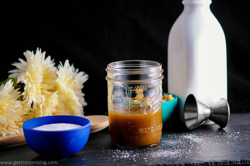 Salted Bourbon Butterscotch Sauce in jar with blue bowl of salt, jigger, white bottle and flowers