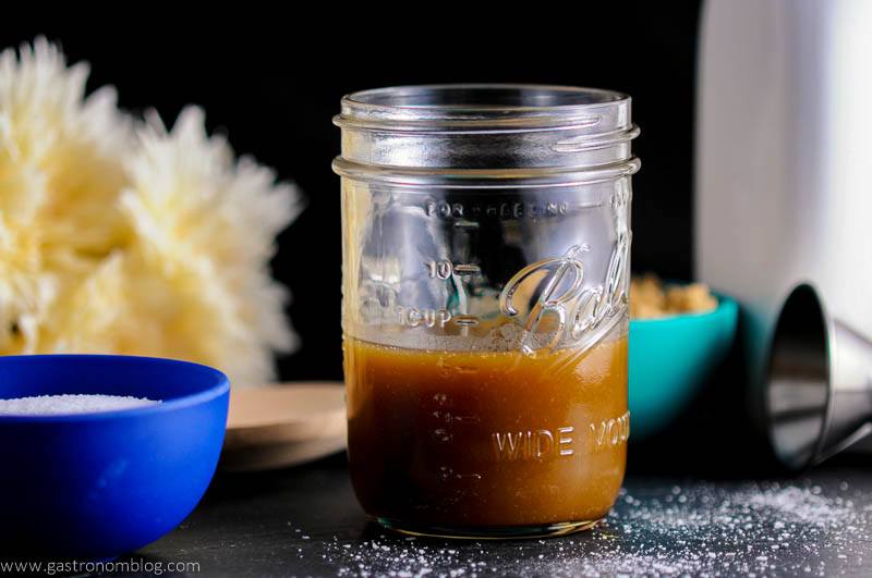 Salted Bourbon Butterscotch Sauce in mason jar, blue bowl of salt, flowers and jigger