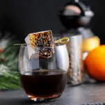 Cocktail in glass with roasted marshmallow in glass. Orange, coffee beans in jar, coffee grinder and greenery in background