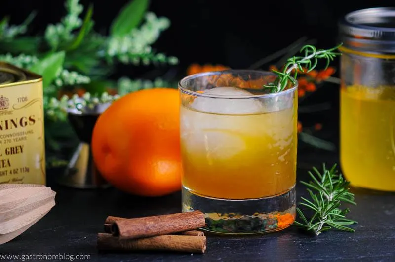 Earl of Orange cocktail, orange cocktail in rocks glass, cinnamon sticks, orange, greenery behind