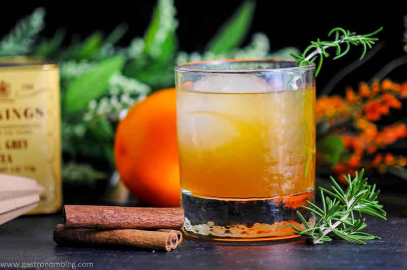 Earl of Orange cocktail in rocks glass with cinnamon sticks and rosemary. Earl Gray tea tin and reamer, orange and flowers