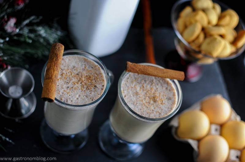 Salted Butterscotch Bourbon Eggnog in glass coffee mugs, cinnamon sticks. Jigger, milk bottle, cookies and eggs in background