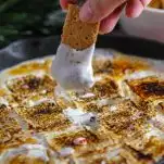 Candy Cane Skillet S'mores with Boozy Marshmallows, hand dipping graham cracker into marshmallows in skillet