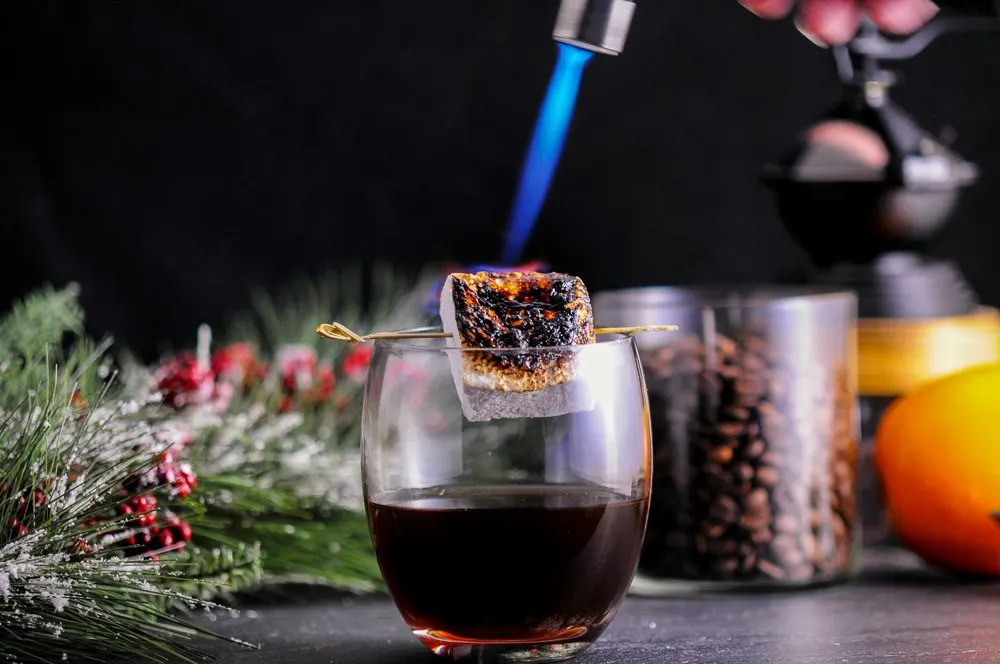 The brown Roast and Toast cocktail with flame on culinary torch for marshmallow. Coffee, fir branch in background