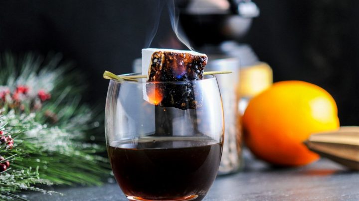 The Roast and Toast, brown cocktail in glass with toasted marshmallow on cocktail pick. Orange, coffee grinder, greenery, coffee beans in jar in background