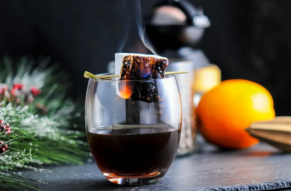 cold brew recipe cocktail in glass with marshmallow on fire. Orange, fir branch and reamer in background
