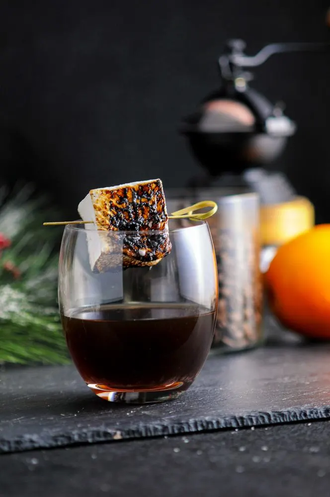 The Roast and Toast, brown cocktail in wine glass with toasted marshmallow on pick. Coffee grinder, beans and orange in background