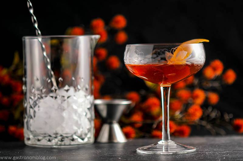 The Woodsman - An Apple Brandy and Ginger Cocktail in a coupe, mixing glass, bar spoon and jigger with flowers in the background