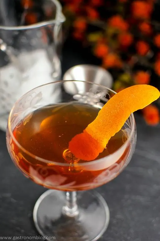 The Woodsman, orange cocktail in coupe with orange peel, mixing glass and orange flowers in background