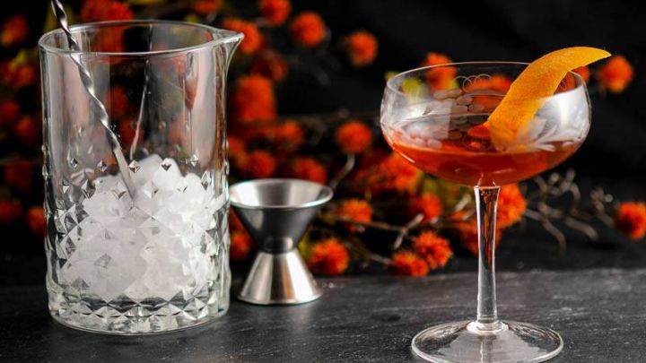 The Woodsman, brown cocktail in coupe with orange peel. Mixing glass with ice, jigger and orange flowers in background