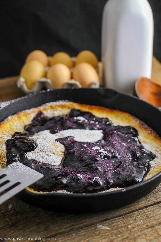 Dutch Baby Pancake with Blueberry Whiskey Sauce in a skillet. Eggs and white jar behind