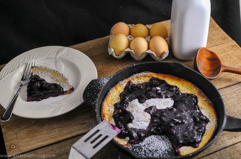 Dutch Baby Pancake with Blueberry Whiskey Sauce in a skillet, some on white plate, eggs, milk bottle and wooden spoon in background