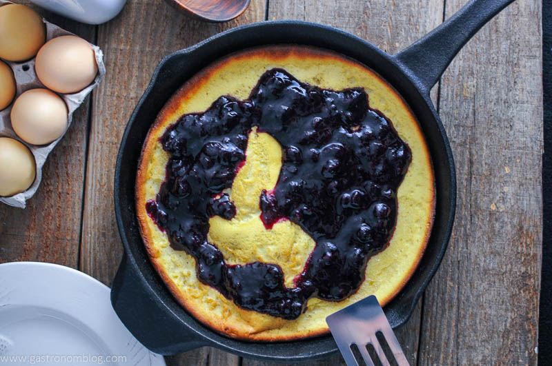 Dutch Baby Pancake with Blueberry Whiskey Sauce in skillet with white plate, eggs and spatula