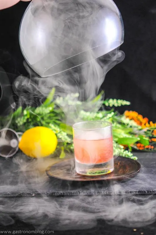 The Smoked Cherry cocktail in rocks glass, cloche over the top with smoke coming out. Lemon and flowers in background