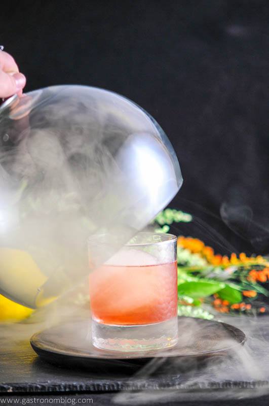 The Smoked Cherry, pink cocktail in rocks glass, smoke and under cloche smoke, flowers in background