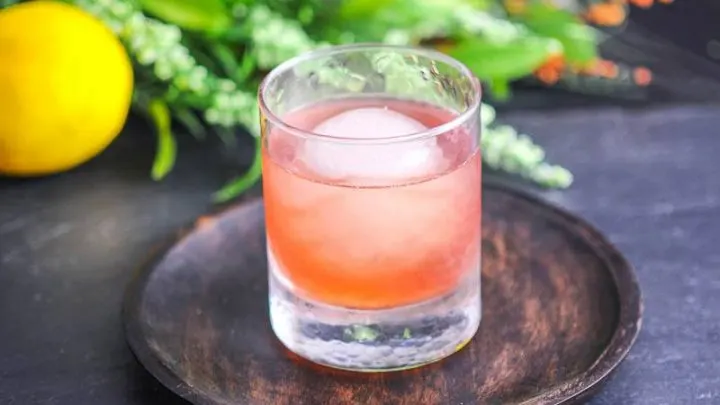 The Smoked Cherry, pink cocktail in rocks glass with ice ball on wooden tray. Greenery and lemon in background