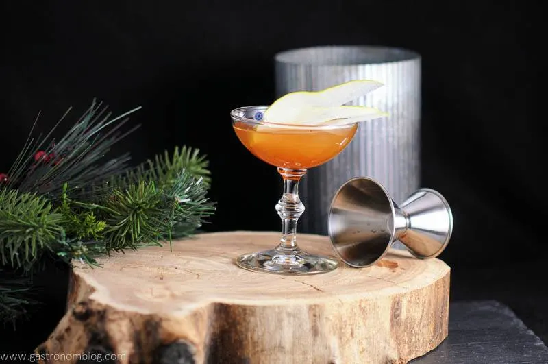 The Peaty Pera cocktail in coupe on wood plate. Jigger, metal canister, fir branch in backgrund