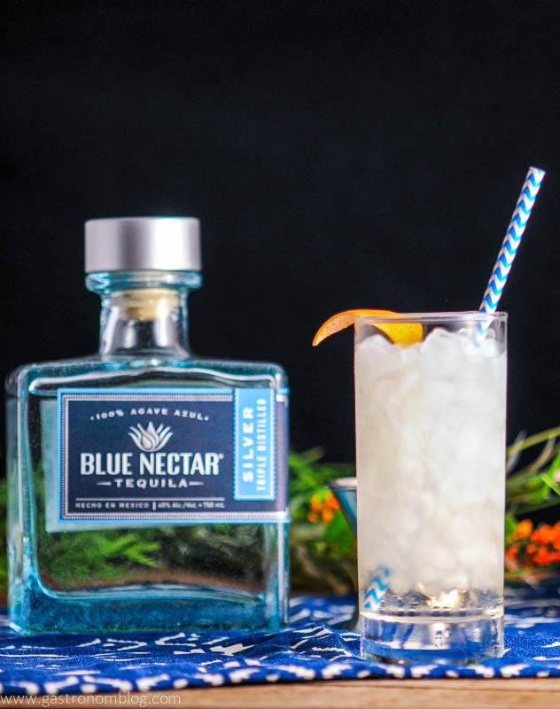 Where The Sea Meets the Sky in a highball with blue straw, citrus peel. Blue tequila bottle on blue and white napkin