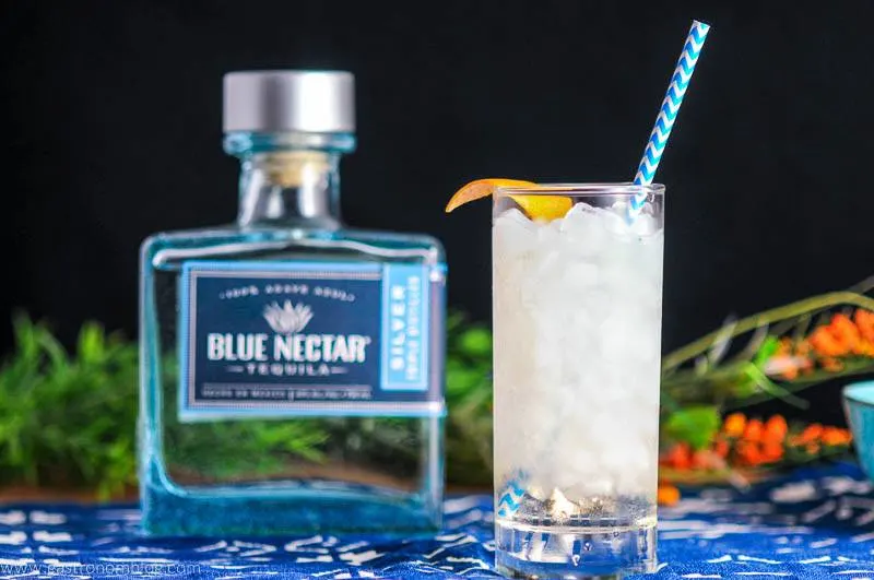 Where The Sea Meets the Sky in higball glass with blue straw and grapefruit twist. Blue tequila bottle and flowers in background