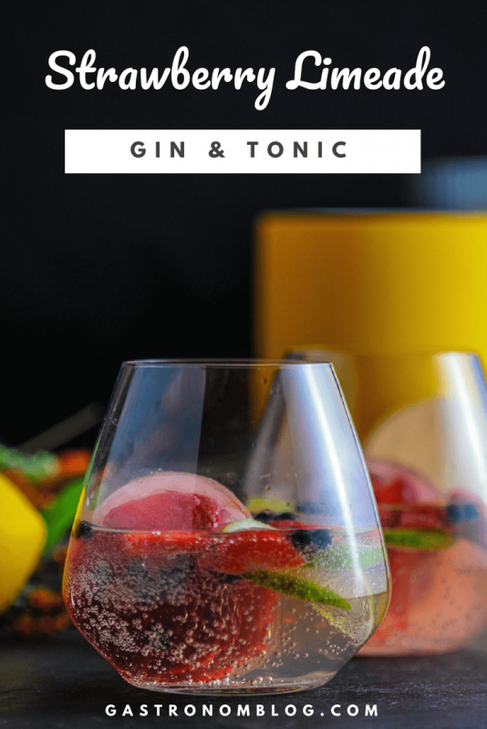 Strawberry Limeade Gin and Tonic in a glass with strawberry ice cube, berries, limes and juniper berries. Lemons, yellow canister and flowers in background