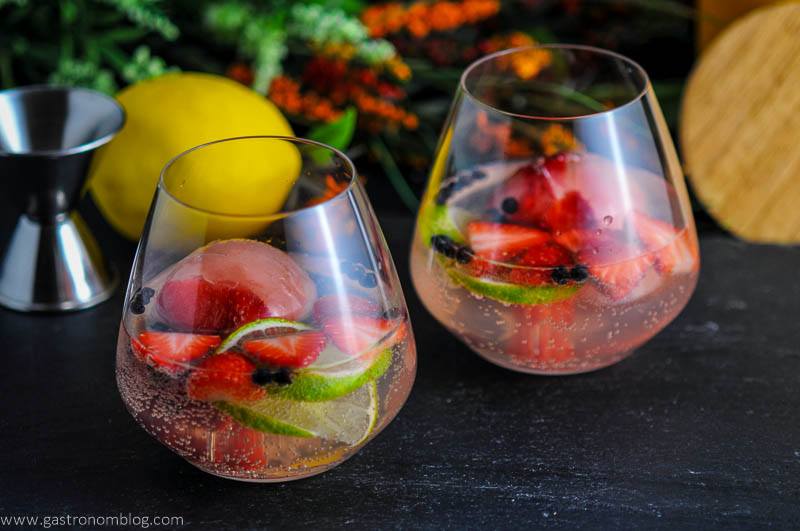 Strawberry Limeade Gin and Tonic Cocktails in glasses with strawberries, limes and juniper berries. Jigger, lemon and flowers in background