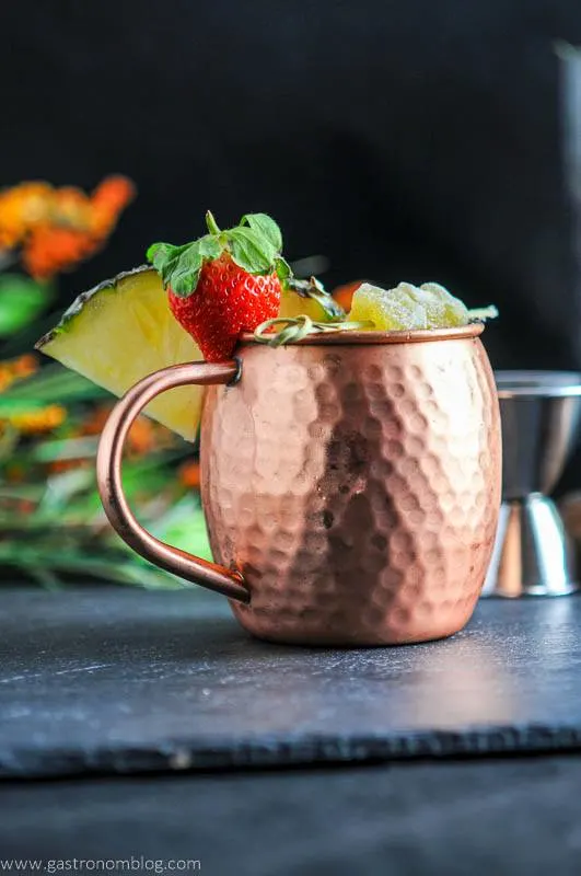 Spicy Pineapple Strawberry Moscow Mule in copper mug with ginger, strawberries and pineapple wedge. Jigger in background
