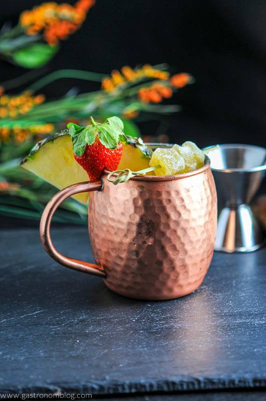 Spicy Pineapple Strawberry Moscow Mule in copper mug with pineapple slice and strawberry. Flowers behind with silver jigger.