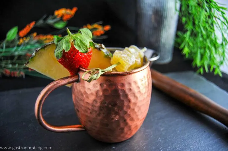 Spicy Pineapple Strawberry Moscow Mule in copper mug. With ginger, strawberry, pineapple wedge. Flowers and muddler in background