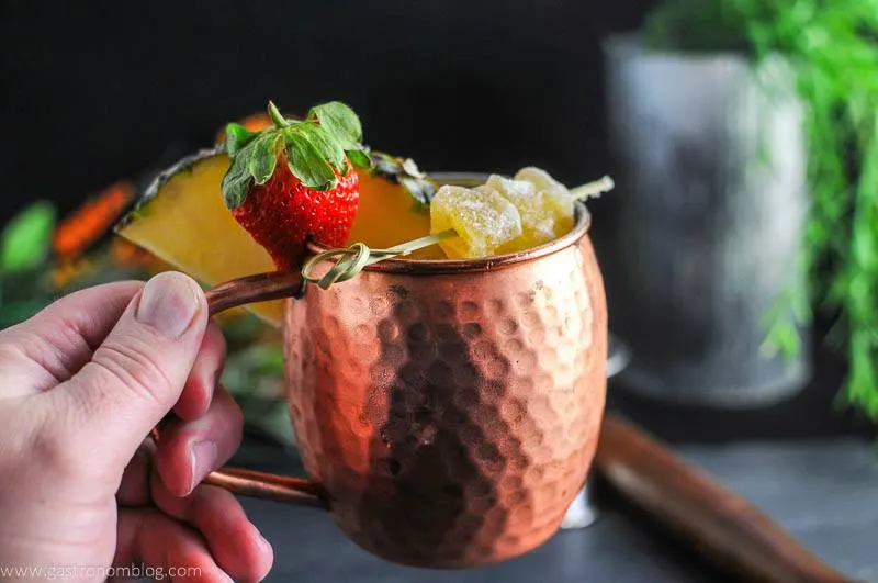 Spicy Pineapple Strawberry Moscow Mule in copper mug, strawberry and pineapple wedge. Crystalized ginger on pick. Held by a hand
