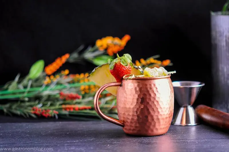 Spicy Pineapple Strawberry Moscow Mule in copper mug with ginger, pineapple wedge and strawberries. Jigger and flowers in background