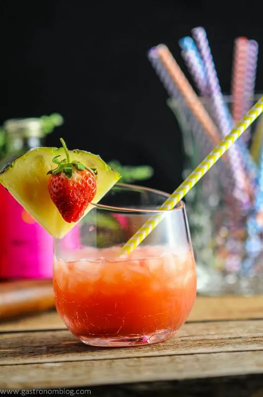 The Nene Bird - A Rum and Campari Cocktail in glass with yellow straw, pineapple and strawberry. Straws in background