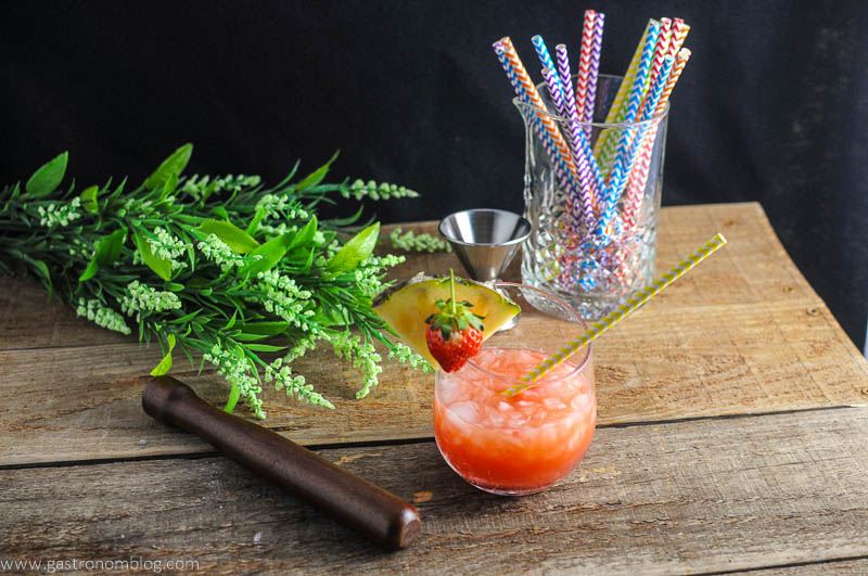 The Nene Bird - A Rum and Campari Cocktail in glass with muddler and flowers, jigger and straws in background
