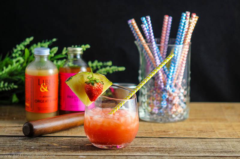 The Nene Bird - A Rum and Campari Cocktail in glass. Yellow straw, pineapple slice and strawberries. In background there is Liber and Co bottles, straws and muddler. 