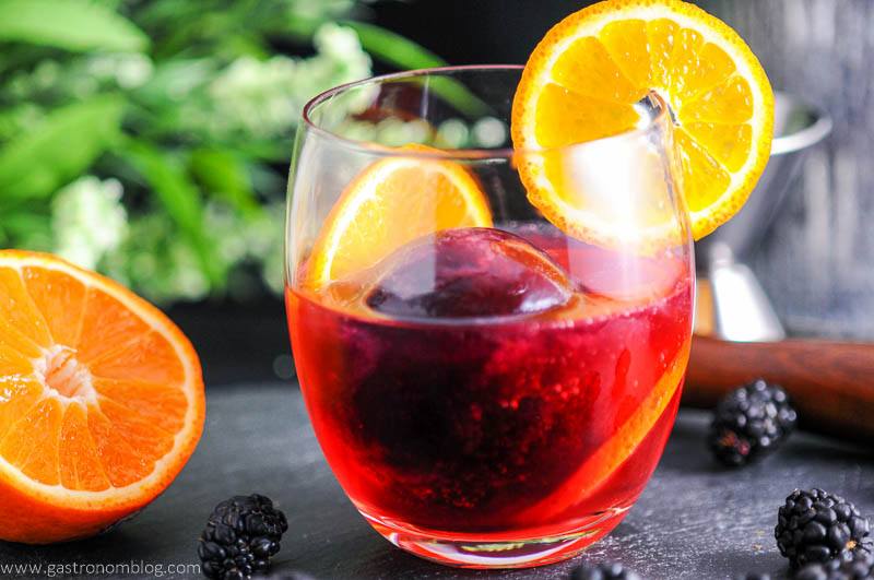 Blackberry Tangerine Vodka Tonic Cocktail in glass with orange wheel, blackberries, orange half and flowers in background