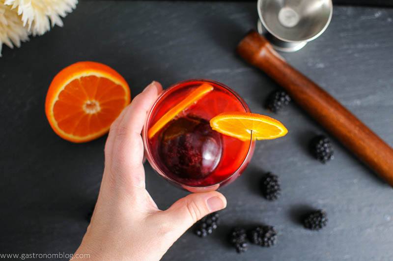 Blackberry Tangerine Vodka Tonic Cocktail in glass, hand holding it. Orange half, blackberries, muddler and jigger